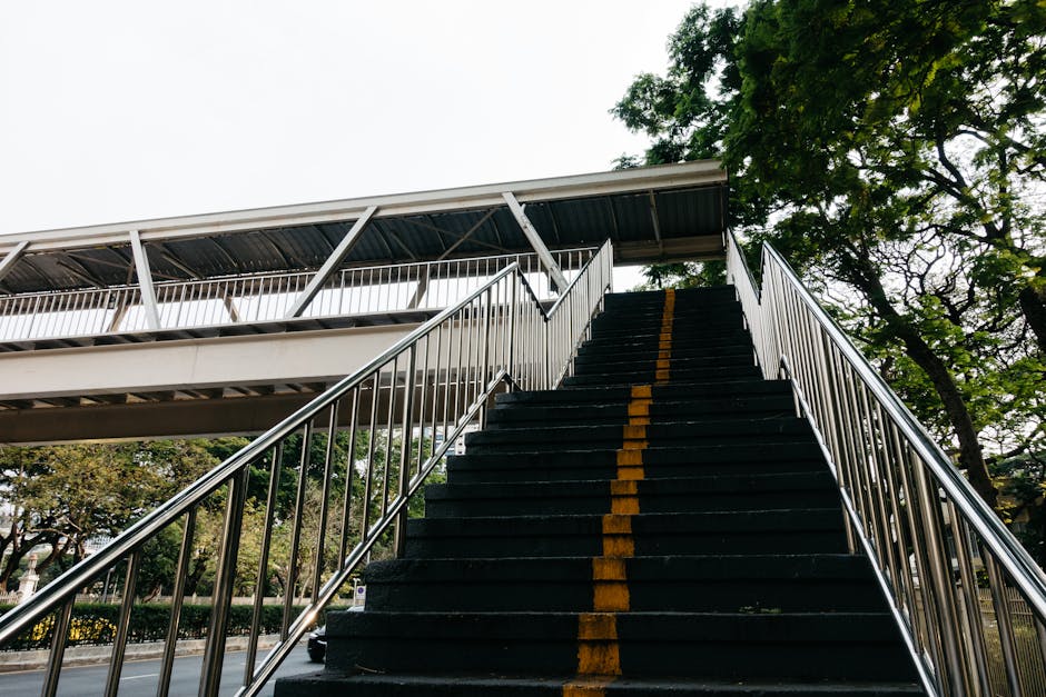 A set of outdoor staircase with black steps marked by a yellow line down the center, leading up to an elevated pedestrian walkway with a metal roof supported by brackets. The staircase has silver metal handrails on both sides. Surrounding the staircase are tall green trees with dense foliage, and the sky above is overcast. This scene illustrates a typical access point at a residential or public property, relevant for home relocation or moving services provided by Man with Van Botany Bay, showcasing the logistical considerations of staircases and outdoor access during furniture transport or packing and moving processes.