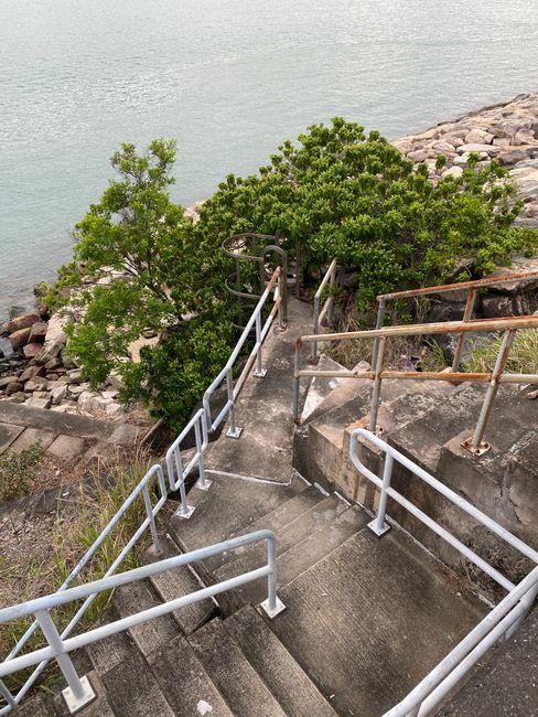 A concrete staircase with metal handrails descending towards a rocky shoreline and calm water, situated outside a building with accessible features including a circular metal lift at the top of the stairs. The staircase is surrounded by greenery and is positioned on a sloped area, indicating challenges with access that may require alternative solutions for home relocation or furniture transport. This scene illustrates the need for careful planning during house removals at locations with staircase and parking access problems, as managed by Man with Van Botany Bay, a professional removals service.