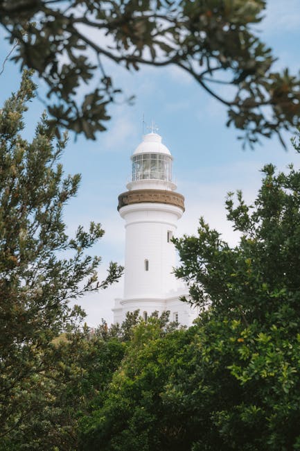 A tall white lighthouse with a cylindrical shape and a balcony near the top, featuring a glass-enclosed lantern room with a domed roof, is visible rising above dense green foliage of trees and shrubs, set against a partly cloudy sky. The scene captures the lighthouse's prominent position on a shoreline, framed by branches and leaves in the foreground. This setting relates to house removals and relocation services by illustrating the importance of careful planning in transporting furniture and equipment, with the lighthouse indicating a coastal area such as Botany Bay. Man with Van Botany Bay’s moving process may involve navigating narrow streets and parking tips, as suggested by the lighthouse’s seaside location and surrounding environment, emphasizing the need for professional loading, packing, and transport of household items during home relocation projects.