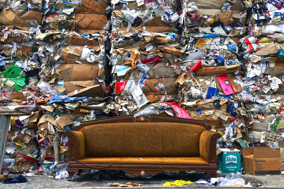 A vintage wooden sofa with fabric upholstery is positioned in front of a large pile of compressed mixed waste materials, including crumpled cardboard, paper, and plastic packaging, which forms a dense wall behind it. The sofa, located on a pavement or outdoor area, appears to be part of a home relocation or disposal process, with various moving and packing materials visible nearby. The background shows a chaos of discarded debris, while the lighting suggests an outdoor setting during the daytime. This scene illustrates furniture disposal or waste clearing, relevant to bulky furniture disposal options for homes in Botany Bay, as managed by Man with Van Botany Bay, a professional removal service specializing in house removals and furniture transport.