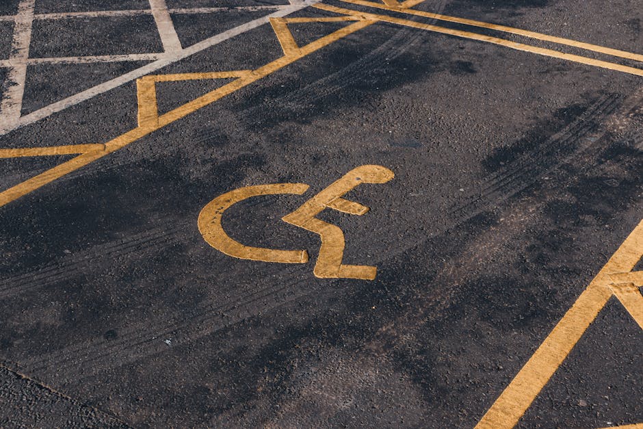 Close-up view of a designated disabled parking space on an asphalt surface, featuring a yellow wheelchair symbol painted on the ground. The parking area is marked with additional yellow lines indicating the boundaries of the space, situated on a paved surface with visible tire marks and slight wear. Adjacent to the parking space, part of the nearby pavement and the edge of the parking lot can be seen. The lighting suggests natural daylight, highlighting the clear, bright yellow paint against the dark asphalt. The image is taken in an urban environment, possibly outside a residential or commercial property, and may be associated with house removals or relocation services provided by Man with Van Botany Bay, with the parking space designated for accessible vehicle access during moving activities.