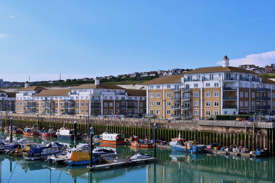 A panoramic daytime view of a marina with numerous small boats docked in calm water, surrounded by wooden piers and mooring poles. Behind the marina, a row of modern, multi-storey residential buildings with white and beige facades, glass balconies, and brown roofs lines the waterfront. The buildings are positioned along a paved walkway, with vehicles parked nearby. In the background, a hillside features additional houses and greenery, under a clear blue sky. The scene reflects a peaceful coastal environment suitable for house removals and home relocation services, with an emphasis on transport and logistics planning by companies like Man with Van Botany Bay.