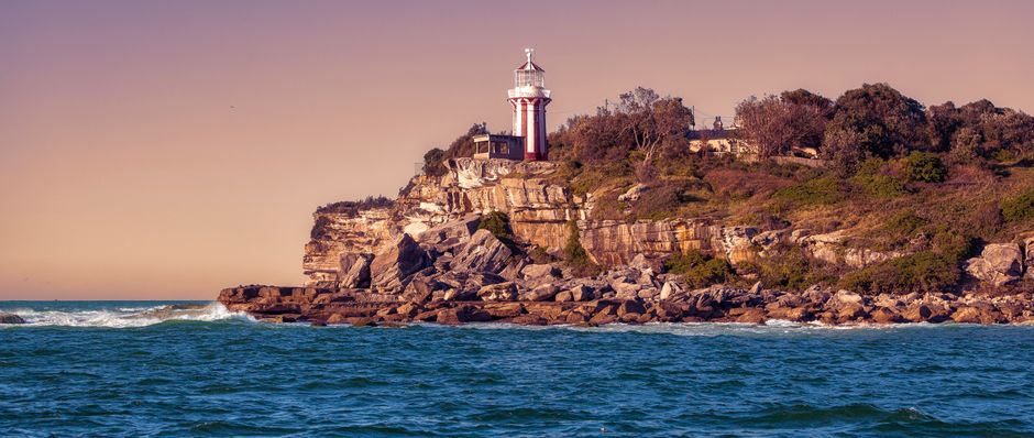 A lighthouse with a red and white tower situated on a rocky cliff overlooking the sea, with several trees and bushes on the hillside behind it. The scene is captured during daylight with clear weather, and gentle waves are visible in the water below. In the foreground, the ocean stretches across the image, displaying blue-green water with some white foam near the rocks. The lighthouse is positioned towards the top of the cliff, providing a visual marker for navigation. The overall setting suggests a coastal area suitable for home relocations involving transport across water or along scenic shorelines, as managed by companies like Man with Van Botany Bay. The image illustrates the natural environment often encountered during a comprehensive furniture transport or moving process.