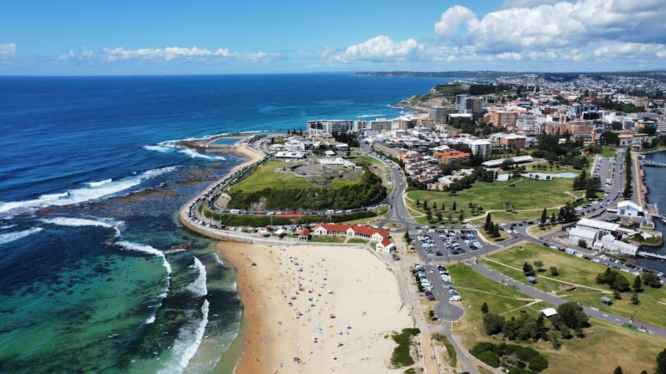 An aerial view of a coastal town showcasing a sandy beach with beach umbrellas and people, adjacent to the shoreline with waves crashing against rocks. Behind the beach, there are green grassy areas and a network of roads with parked cars. The town features a mix of modern buildings, residential houses, and commercial structures. A large park with trees and open spaces is visible near the centre, along with a small marina or port area on the right. The background extends to a cityscape with a variety of multi-storey buildings and the ocean stretching to the horizon under partly cloudy skies. The image illustrates a scenic seaside environment suitable for house relocations and moving logistics, highlighting the urban and coastal surroundings managed by companies like Man with Van Botany Bay for efficient furniture transport and packing services.