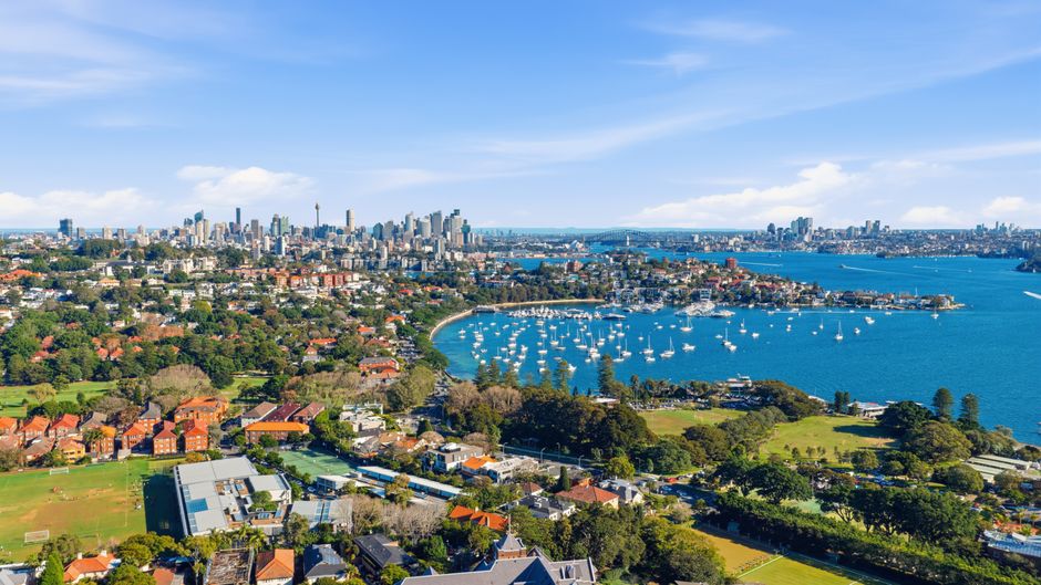 Aerial view of Botany Bay showing a residential area with houses and greenery in the foreground, alongside a marina filled with numerous sailboats on the calm blue water. Beyond the bay, a dense city